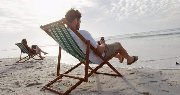 Side view of young caucasian man sitting on sun lounger and using mobile phone at beach 4k
