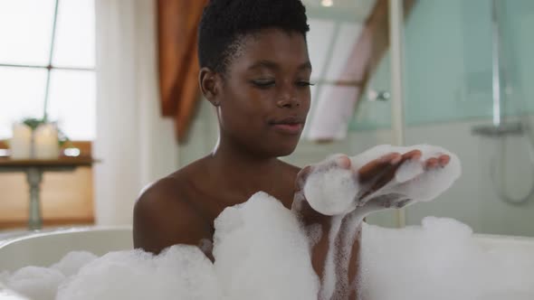 Smiling african american attractive woman taking bath and blowing out foam in bathroom