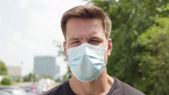 A Young Handsome Man in a Face Mask Looks at the Camera - Closeup - an Urban Area in the Blurry