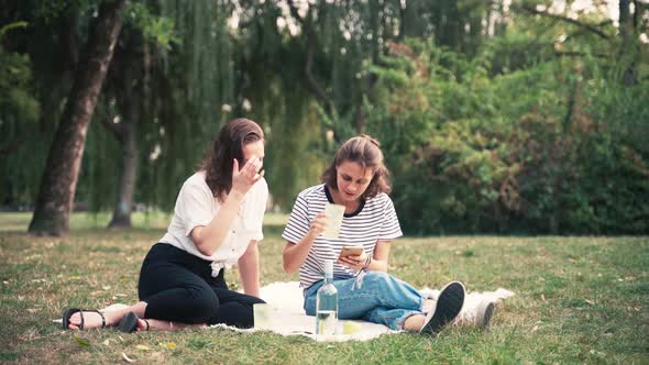 Two Young Women Sitting on a Blanket in the Park and Drinking Wine