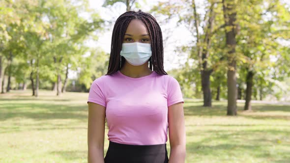 A Young Black Woman in a Face Mask Shows a Thumb Up To the Camera and Nods in a Park on a Sunny Day