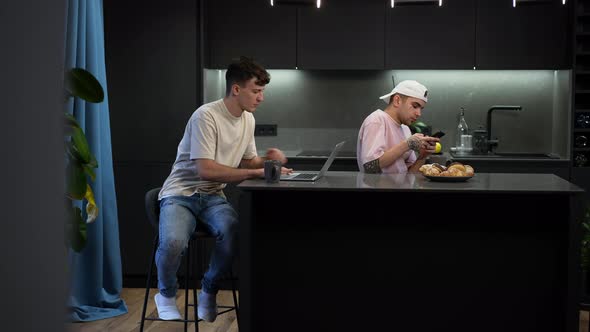 Wide Shot Young Caucasian Gay Couple Messaging Online on Laptop Talking on Phone Sitting in Kitchen