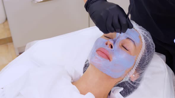 Close-up, Cosmetician in Black Gloves, Applies Blue Cosmetic Face Mask with Special Brush To Female