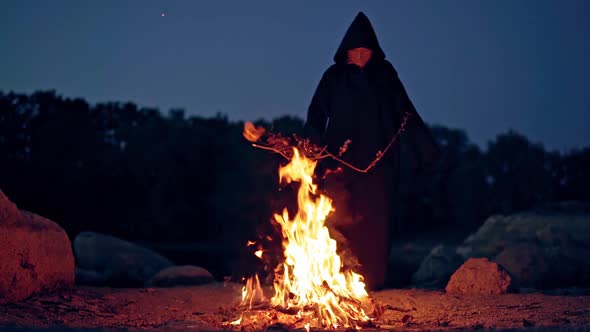 Witch makes scary ritual in the fire. Mysterious wizard in black cloak doing shamanism over the brig