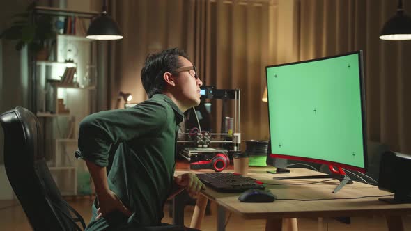 Asian Man Paining His Back While Work On 3D Printer And Computer With Mock Up Green Screen Display