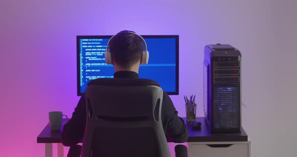 Rear View of Man Programmer in Headphones Coding Network Security Software Indoors in Neon Lights at