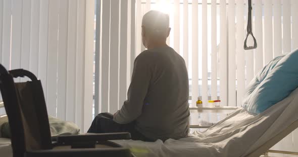 Senior Disabled Man Sitting on Hospital Bed and Looking at Wheelchair
