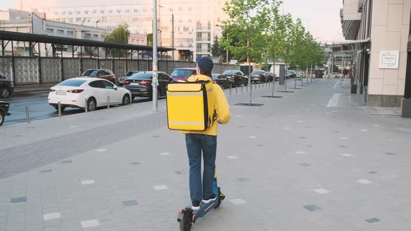 Male Courier with Thermal Backpack Riding Electric Scooter Outdoors Along Urban Building While