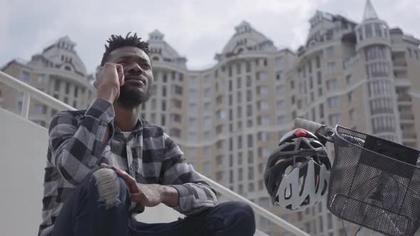 Handsome African American Man Sitting Near His Bicycle on the Background of Urban Architecture. The