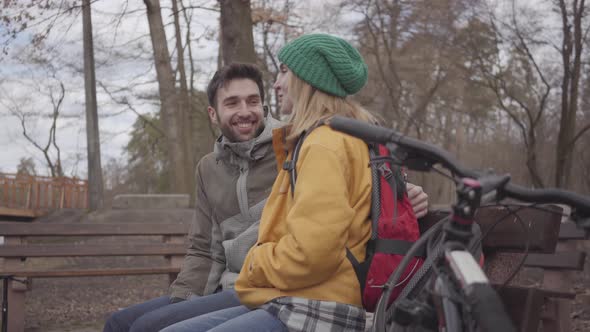 Joyful Couple of Tourists with Backpacks Sitting on the Bench in the Spring Park at the Riverbank