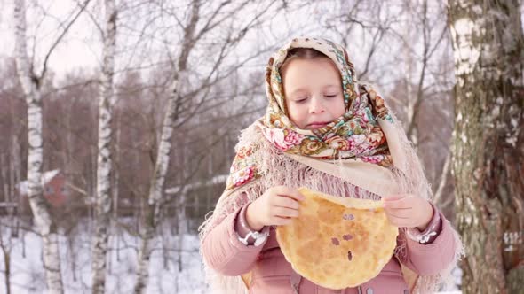 Cute girl in a traditional Russian headscarf with bagels eats pancake on winter background.