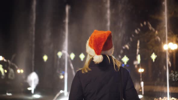 A girl looks at a fountain on Christmas night in a Santa Claus hat