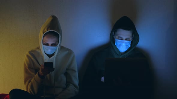 Young Couple in Medical Masks Using Smartphone and Laptop at Night