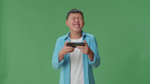 Laughing Young Asian Boy Looking At The Phone Screen On Green Screen In The Studio alt