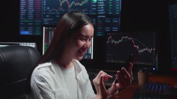 Asian Female  Stock Market Broker Using Mobile Phone With Analyzing Graphs On Multiple Computer