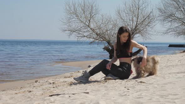 Brunette in Top and Jeans Sits on Sand Sea Beach with Dog