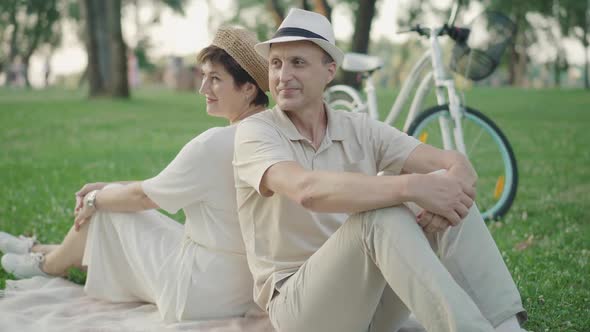 Cheerful Mid-adult Man and Woman Sitting Back To Back on Green Lawn and Talking