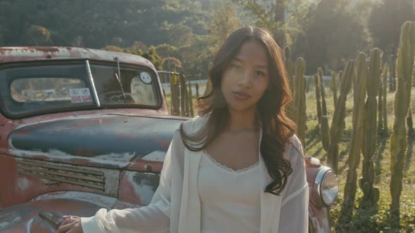 Young Asian Woman Stands By an Old Pickup Truck at Sunset and Looks at the Camera