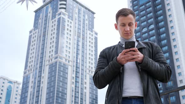 Handsome Man in Leather Jacket with Phone on Background of Tall Houses