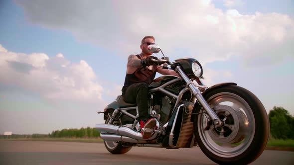 Stylish Biker with Tattoos Rides a Motorcycle on a Country Road at Sunset