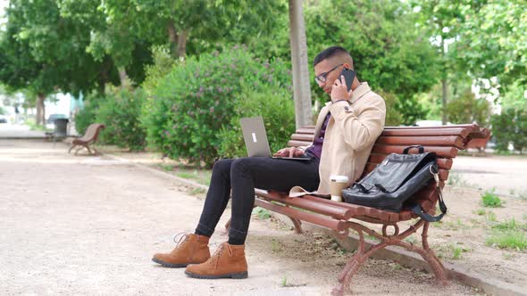 Young businessman in trendy outfit speaking on phone on bench