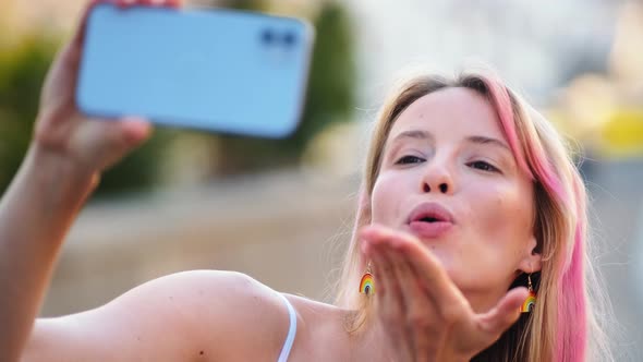 A smiling woman with rainbow earrings sending an air-kiss using her phone camera during pride gay pa