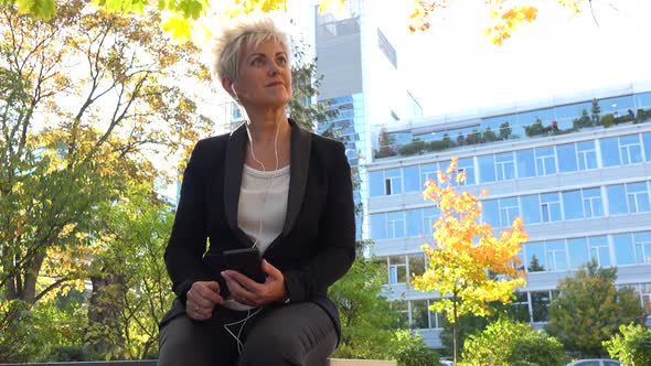 A Middle-aged Businesswoman Sits on a Bench in a Park and Listens To Music on a Smartphone