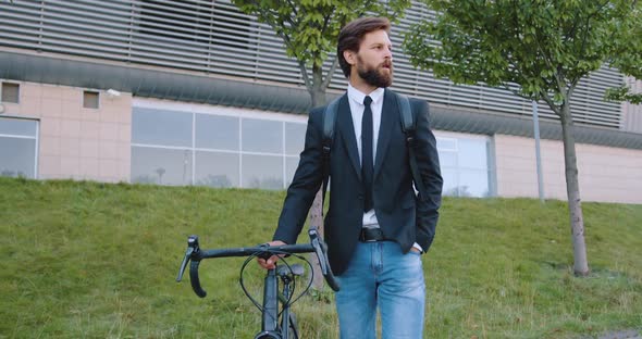 Bearded Man in Dark Jacket and Jeans which Goes with His Bike on the Street