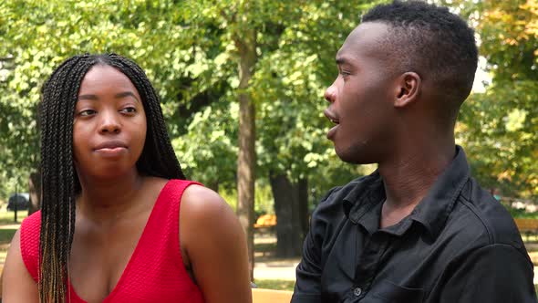 A Black Man and a Black Woman Sit on a Bench and Talk in a Park on a Sunny Day - Closeup