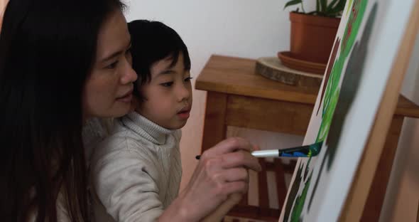 Asian mother having fun doing color paint with little son on canvas at home