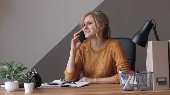 a Young Beautiful Woman of European Appearance Sits in an Office with a Phone and a Work Pad