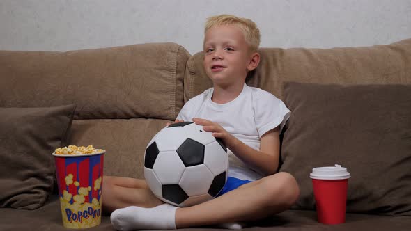Little Boy Is Watching a Football Match on TV Sitting on the Couch with Popcorn