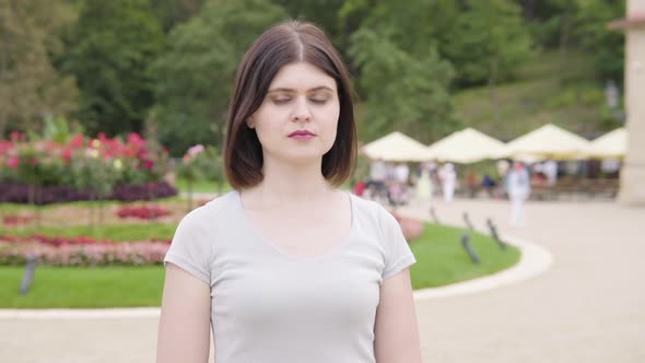 A Young Caucasian Woman Looks Seriously at Camera in a City Park  People at Tables in a Restaurant