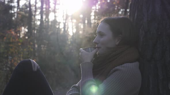 Sun shines over stunning woman in cozy sweater, jeans and boots drinking hot tea in fall forest