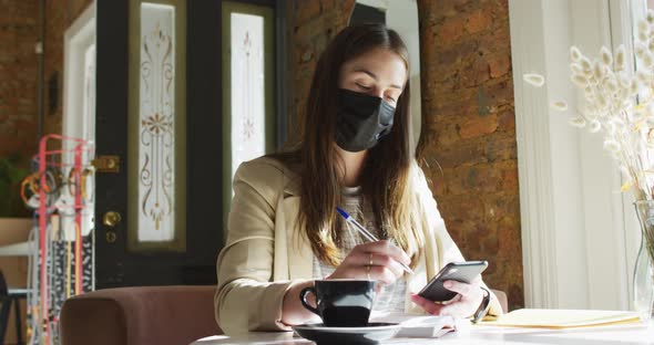 Caucasian female customer wearing face mask, sitting at table, using smartphone