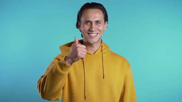 Positive young man smiles to camera. Hipster guy showing thumb up sign over blue 