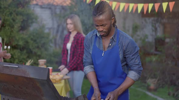 Middle Shot of Focused African American Young Man in Apron Preparing Meat for Barbecue with Blurred