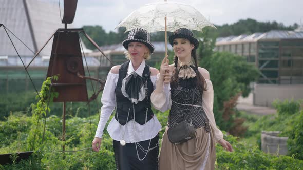 Cheerful Beautiful Mother and Daughter in Steampunk Costumes Strolling with Sun Umbrella Leaving