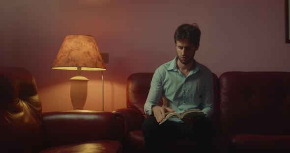 Young guy with shirt reads a book sitting in the living room at home