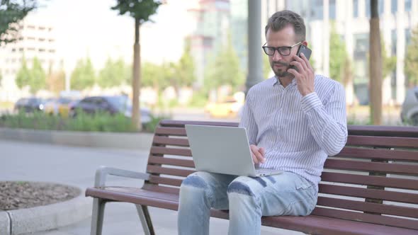 Young Adult Man Talking on Phone and Using Laptop While Sitting Outdoor on Bench