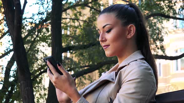 Young Beautiful Woman Sits on the Bench in Park and Works on the Smartphone