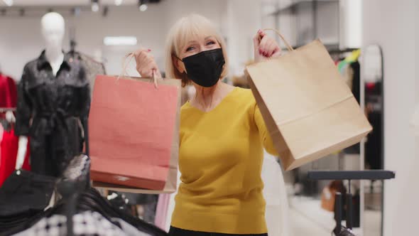 Happy Middleaged Woman in Protective Mask Standing in Clothing Store Holding Bags in Hands