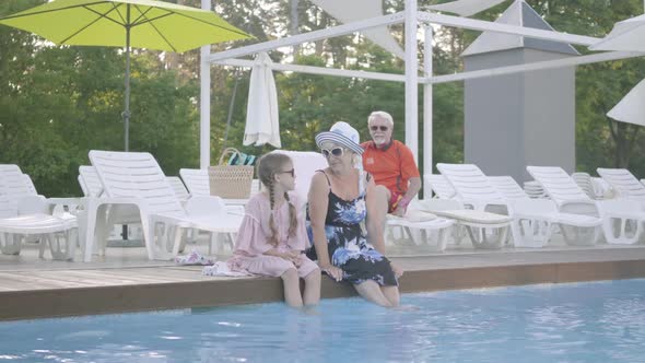 Little Cute Girl with Pigtails and Mature Woman Sitting on the Edge of the Pool with Their Feet