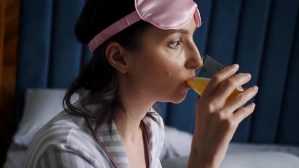 Woman in Striped Pajamas and with a Sleep Mask is Sitting on the Bed in the Bedroom and Drinking