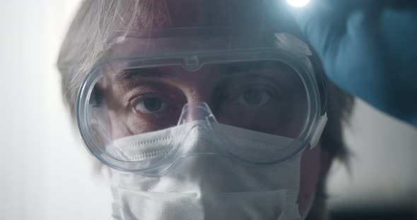 Doctor in Protective Mask and Goggles Testing Eye Reflexes of Patient Using Lamp in Medical Clinic