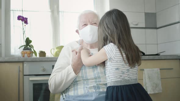 Grandmother Hugs Her Granddaughter Sitting In The Kitchen. They Are Both In Medical Masks.