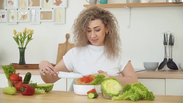 Young Blonde Woman with Curly Hair Throws Sliced Red Tomato Into a Bowl While Sitting at a Table in