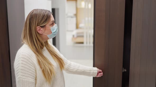 a Young Woman in a Protective Mask in a Furniture Store Chooses a Cabinet Opening the Doors