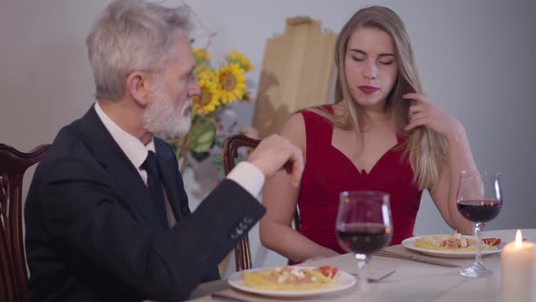 Portrait of Young Seductive Woman in Red Dress Sitting with Senior Man at the Table and Smiling