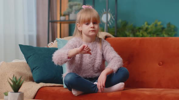 Child Girl Kid Sitting on Sofa at Home Alone Showing Thumbs Down Sign Gesture Dislike Disapproval